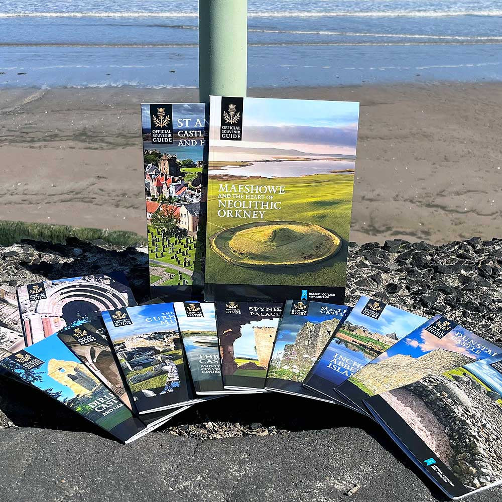 A selection of Historic Scotland guidebooks are displayed on a rocky wall by a beach. Blue waves are in the distance. 