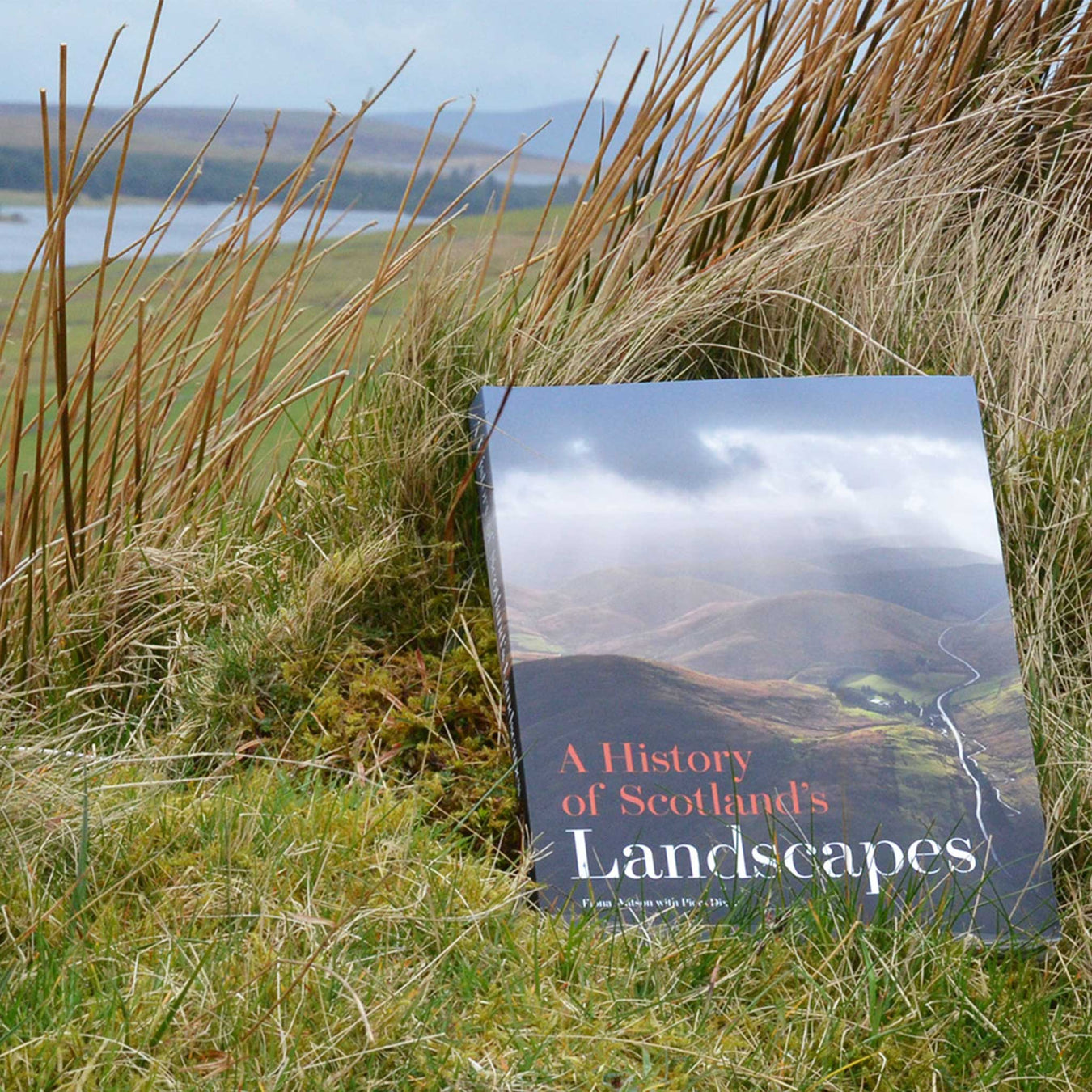 Large paperback copy of our Historic Scotland publication, 'A History of Scotland's Landscapes' lies on a grassy hill looking over a loch. 