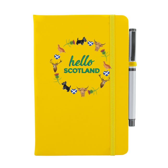 A beautiful designed yellow notebook with the words 'Hello Scotland' in green,  featuring elements of Scottish culture and scenery. A yellow strap closes over the right hand side with a matching pen.