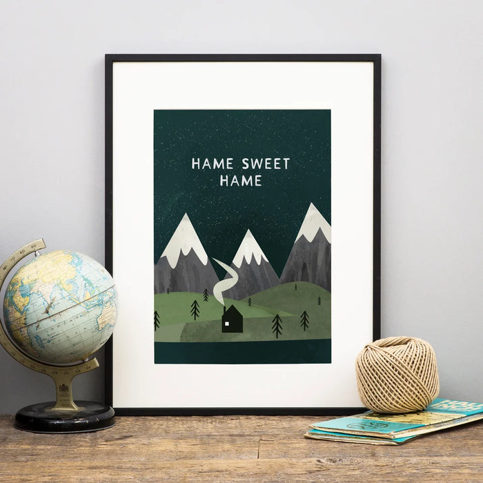 'Hame Sweet Hame' print features a mountain scene with a cottage under a starry night. The digital style image is framed and sitting on a shelf next to a globe and map. 