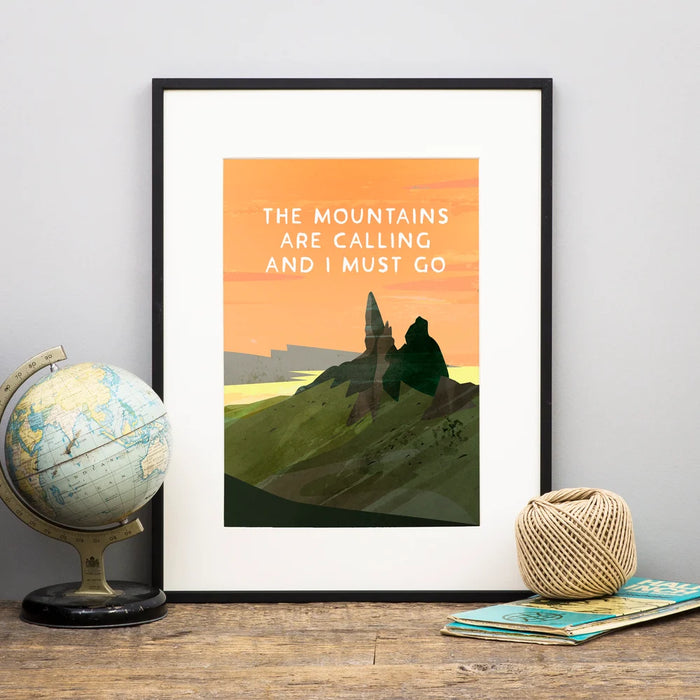 'The Mountains are Calling and I Must Go' digital style print shows a mountain peak against a golden orange sunrise. The framed print is placed on a wooden shelf next to a globe and map. 