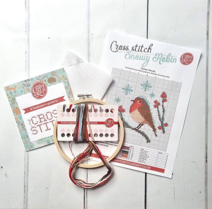 Brightly coloured thread, a hoop and cross stitch instructions for the Snowy Robin Cross Stitch kit lay on a white wooden floor