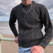 Person leans on a blue seafront barrier wearing the Charcoal Grey Aran Jumper. 