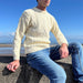 Person sits on a wall in front of the beach wearing a cream Arran knit Jumper 