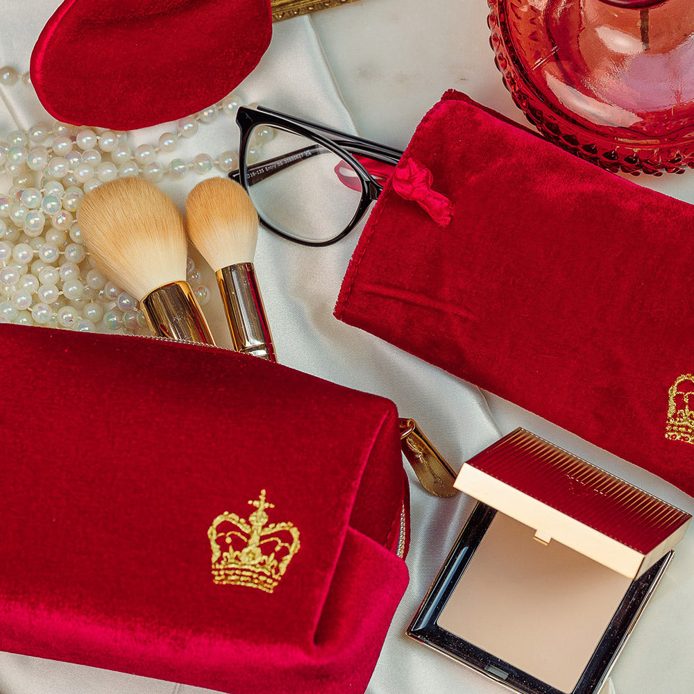 Collection of red velvet accessories including a wash bag and soft glasses case are arranged on a table with pearls, make up brushes and dried flowers. 