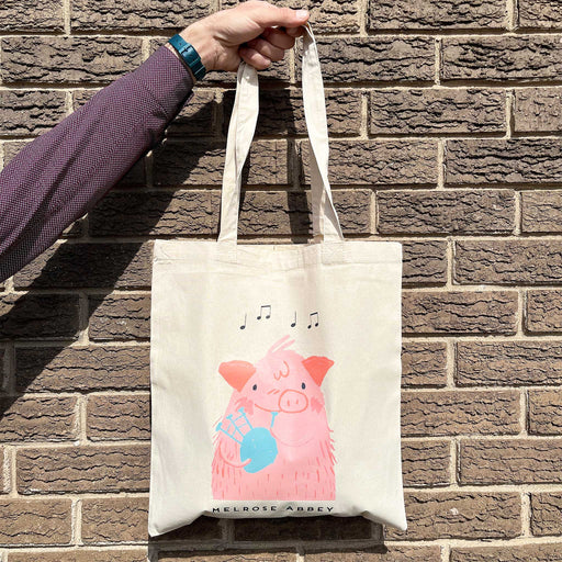 A cotton tote bag is held against a brick wall by a person wearing a purple shirt, only their arm is visible. The tote bag features a print of our Bagpiping Pig from Melrose Abbey. 