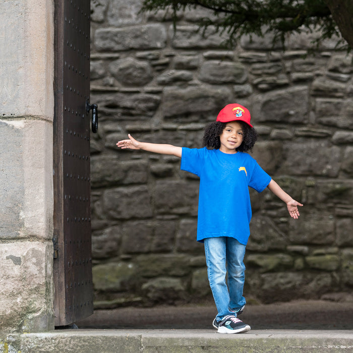 Small child stands with arms outstretched in a castle doorway. They are wearing the blue knight t-shirt and the red knight cap. 