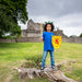 Child stands in front of castle ruins wearing the blue knight Tee. They are holding a wooden sword, a lion rampant shield and are wearing a green dragon head piece. 