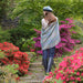Person stands looking over their left shoulder amongst a floral path. The person wears the Bracken Tartan Tam & Fine Scarf. 