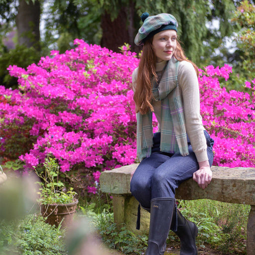 Person sits on a bench next to a large pink floral bush. The person looks to the left and wears the Bracken Tartan Tam & Fine Scarf. 