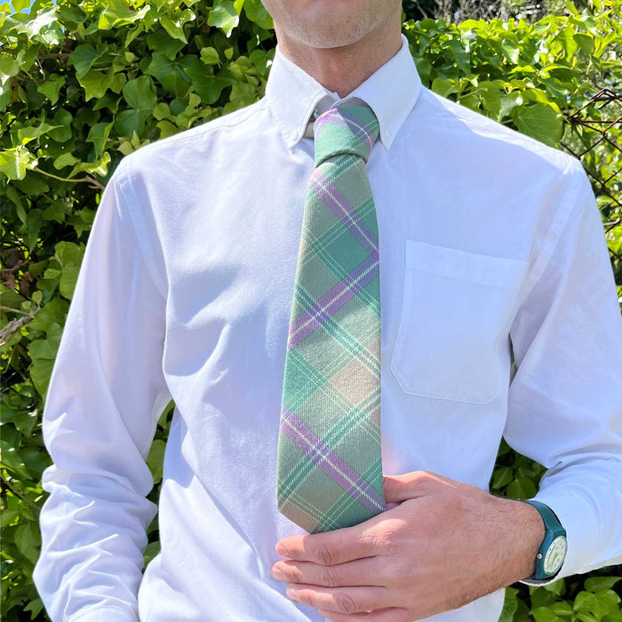 Person stands in front of some greenery wearing a white shirt with the Bracken Tartan Tie. 