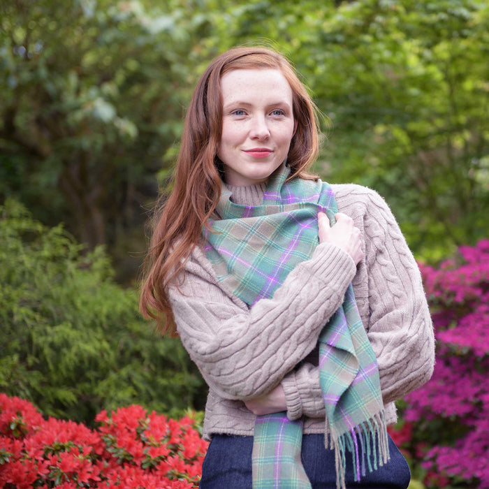 Person surrounded by trees and brightly coloured floral bushes wraps themselves in the Bracken Tartan Wool Scarf.