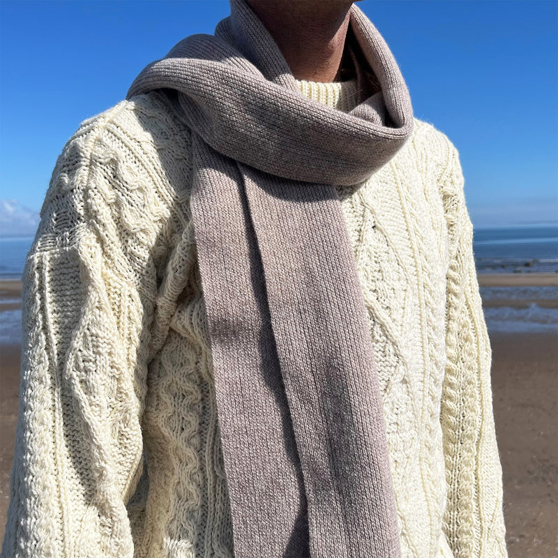 Person stands in front of a sea scene wearing a cream Aran jumper and the beige Castle Cosies scarf.