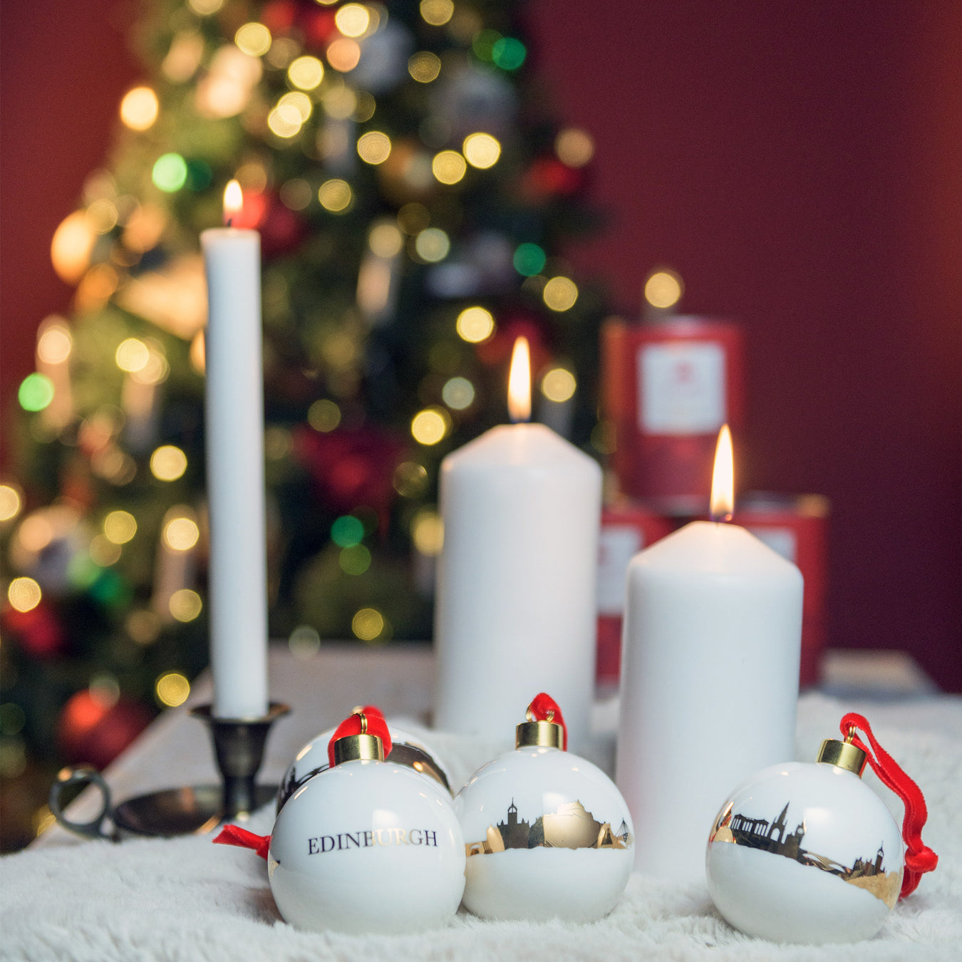 White pillar candles and delicate white baubles with gold details are placed on a fur rig in front of a lit Christmas Tree. 