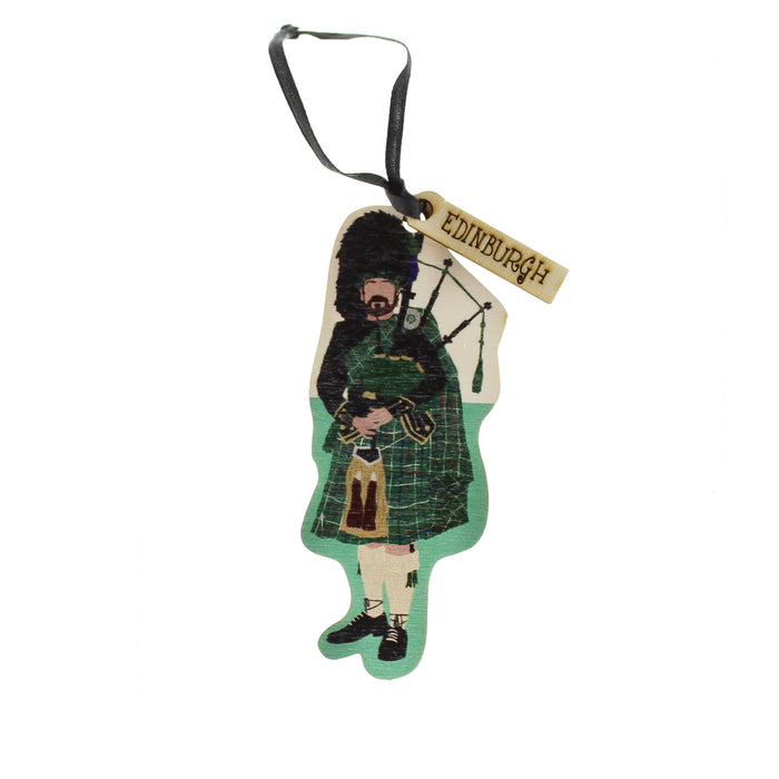 A traditional wooden Christmas decoration depicting a Scottish Piper. The decoration also features a small ribbon and a tag with text that reads ,Edinburgh'.