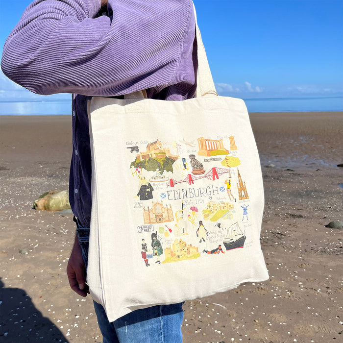 Person wears the Edinburgh Cityscape Tote over their shoulder whilst standing in front of a beach and blue sky. The tote features some of Edinburgh's most loved landmarks. 