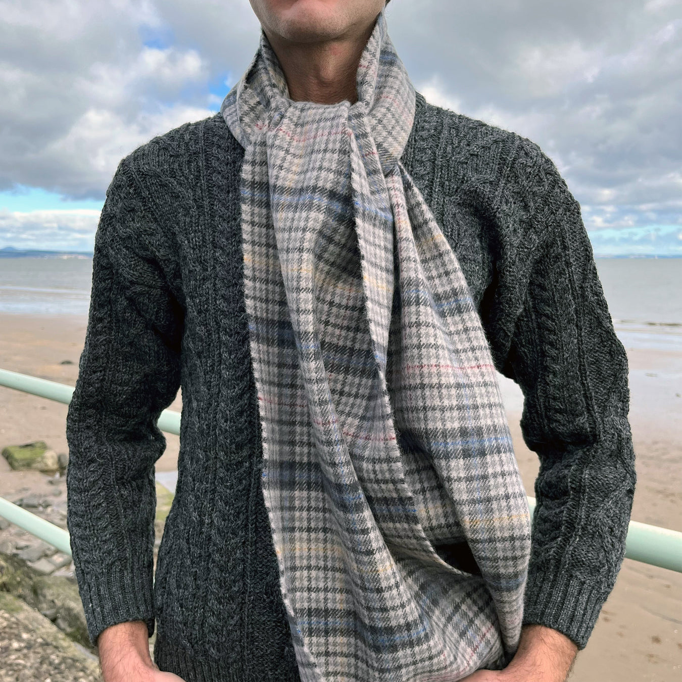 Person stands in front of a beach wearing the Edinburgh Castle Tweed scarf with the charcoal grey Aran Jumper. - Image