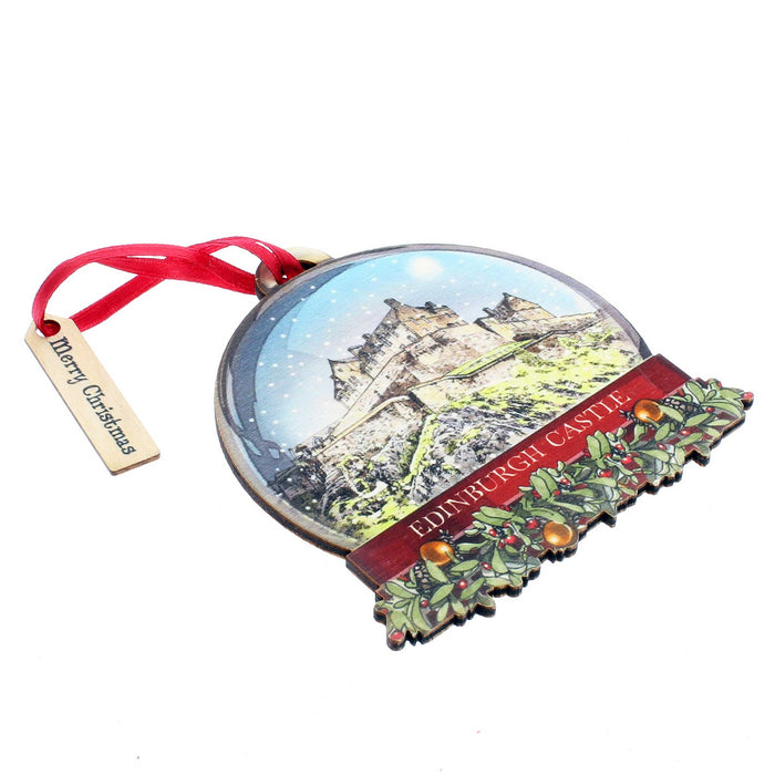 A wooden tree decoration in the shape of a snowglobe. Edinburgh Castle is featured in a snowy scene with holly and festive wreathes at the bottom.  The decoration is laid flat. 