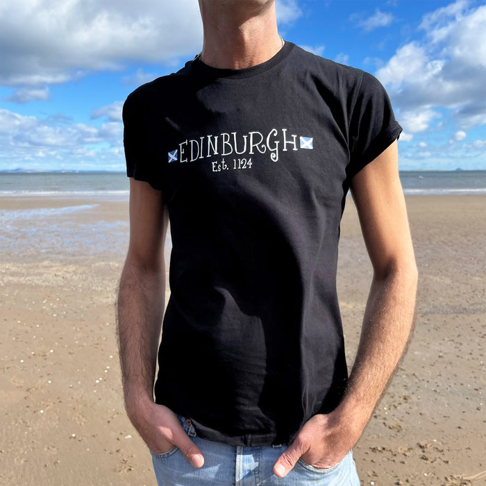 Person wears a black t-shirt with a blue sky and beach scene in the background. The T-shirt has a white print reading Edinburgh, with a St Andrew's flag on wither side. Underneath the Edinburgh there is further details reading Est. 1124, in reference to the age of Edinburgh. 