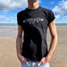 Person wears a black t-shirt with a blue sky and beach scene in the background. The T-shirt has a white print reading Edinburgh, with a St Andrew's flag on wither side. Underneath the Edinburgh there is further details reading Est. 1124, in reference to the age of Edinburgh. 