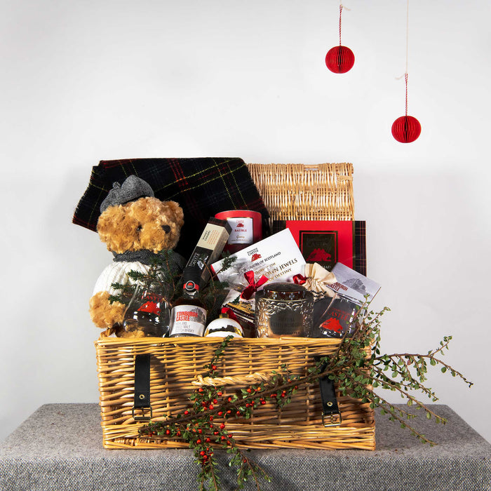 Large wicker hamper sitting on a table features bespoke Edinburgh Castle items including Whisky, chocolates, tea, tartan scarf, shortbread, fudge, whisky accessories and a teddy. 