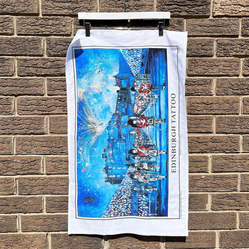 White tea towel hanging against a brick wall features a blue image of the Edinburgh Tattoo in front of the castle.