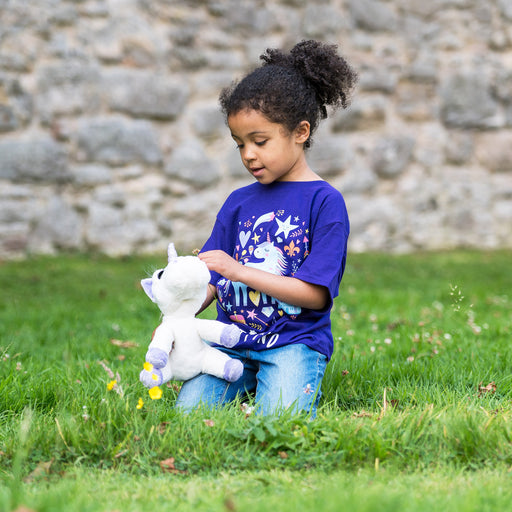 Child plays on the grass with a Unicorn soft toy. They are wearing a purple t-shirt with an enchanted unicorn design.