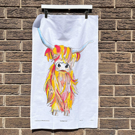 White cotton tea towel hanging on a brick wall features a brightly coloured print of a Highland Cow. 