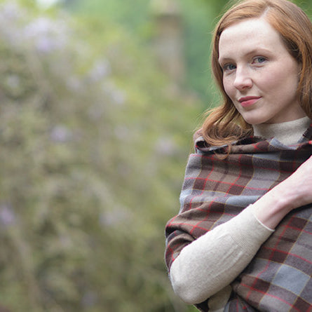 Person wraps themselves in the Outlander tartan serape. The background of a brick wall covered in moss is mostly blurred out.  - Image