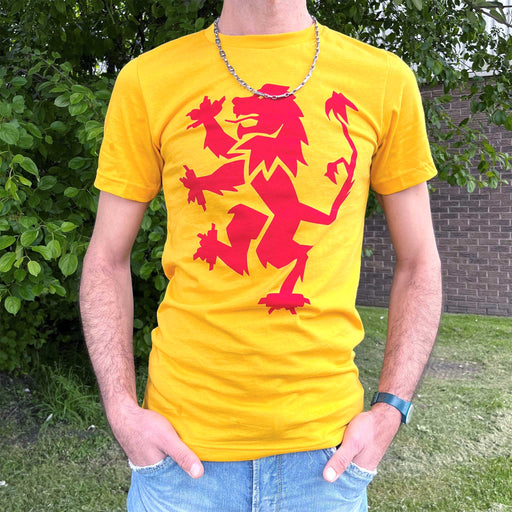 Person standing next to a wall and some greenery wears blue jeans and a yellow t-shirt. The t-shirt features a bold red print of the Scottish Lion Rampant.