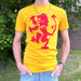 Person standing next to a wall and some greenery wears blue jeans and a yellow t-shirt. The t-shirt features a bold red print of the Scottish Lion Rampant.