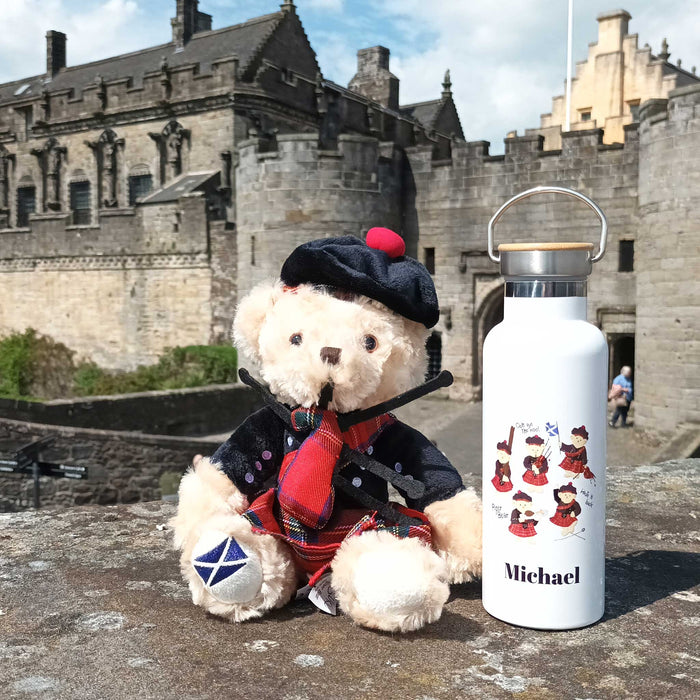 Piper bear sits on a wall in front of Stirling Castle. A white piper bear printed water-bottle sits next to the teddy. 