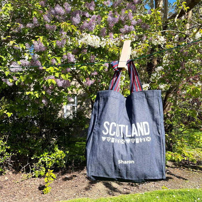 Blue denim tote is hanging on a washing line on front of a purple flowered bush. The tote reads 'SCOTLAND' in white. 