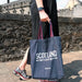 A person walks past a brick wall holding the blue denim Scotland tote bag. 