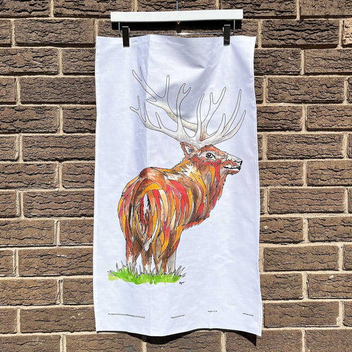 White cotton tea towel hanging on a brick wall. The tea towel features a brightly coloured print of a stag; the monarch of the glen. 