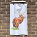 White cotton tea towel hanging on a brick wall. The tea towel features a brightly coloured print of a stag; the monarch of the glen. 