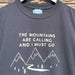 Close up of a black t-shirt hanging on a white hanger against a brick wall. The T-Shirt features a mountain print with the words 'The Mountains are Calling and I must go' printed above.