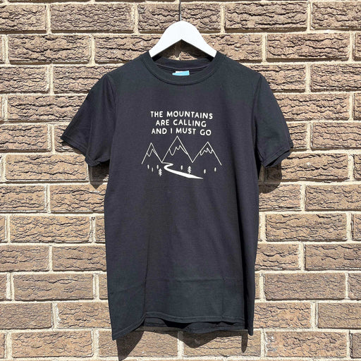 A black t-shirt hanging on a white hanger against a brick wall. The T-Shirt features a mountain print with the words 'The Mountains are Calling and I must go' printed above. 