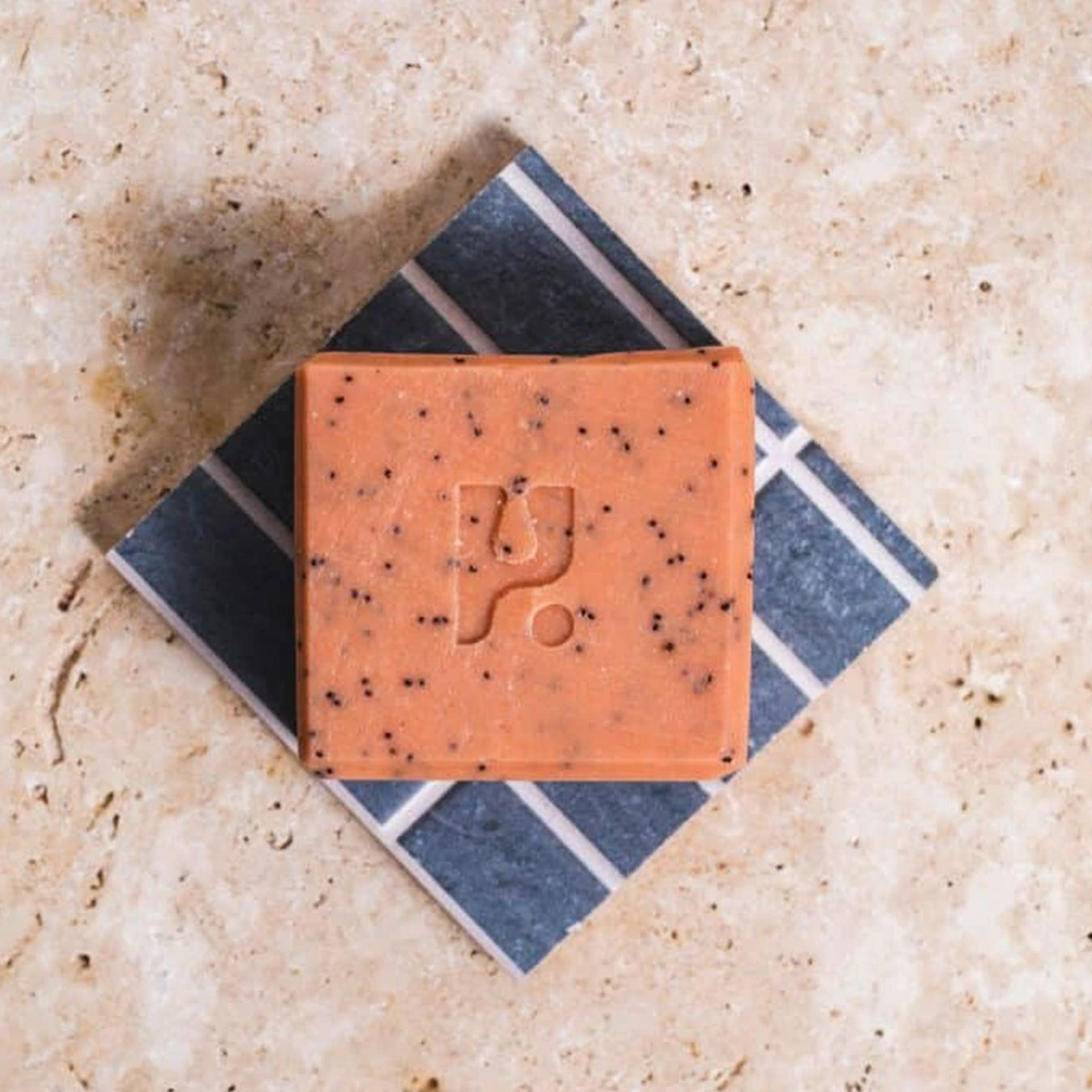 A square bar of rust coloured soap sits on a blue tile and a clay floor. 