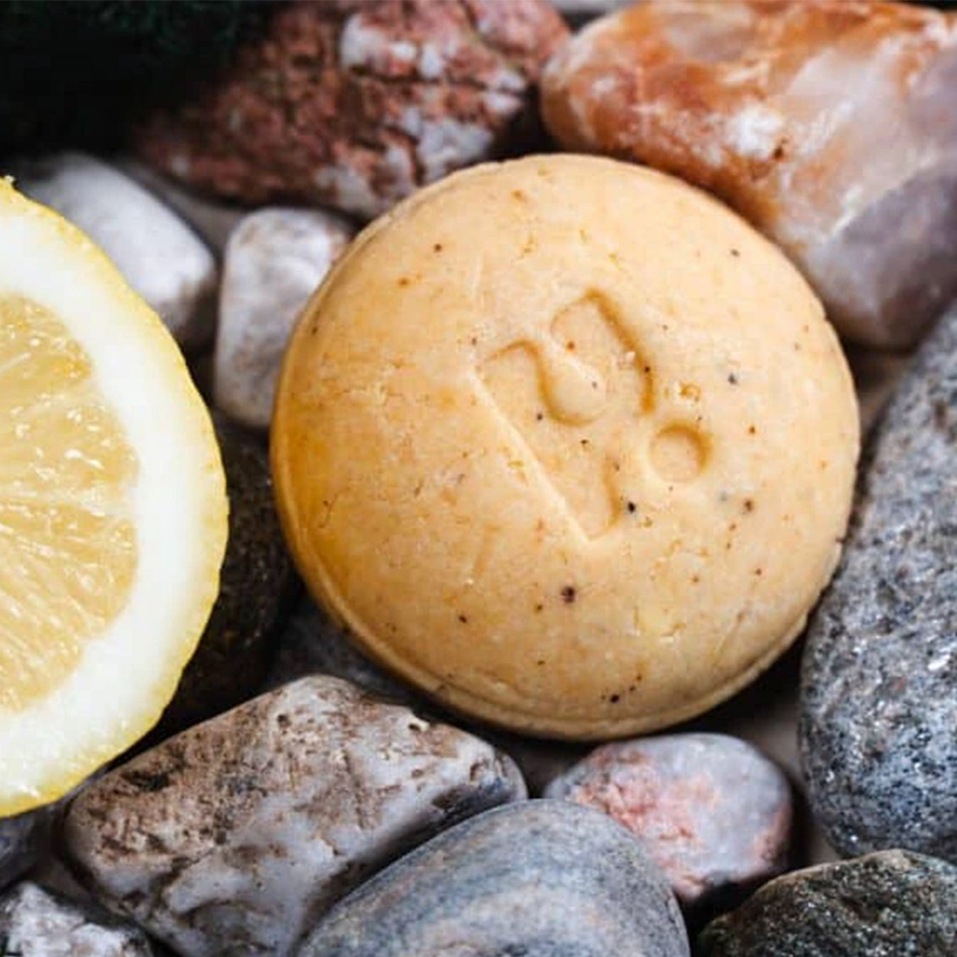 A round yellow soap bar sits among some pebbles and a piece of lemon. 