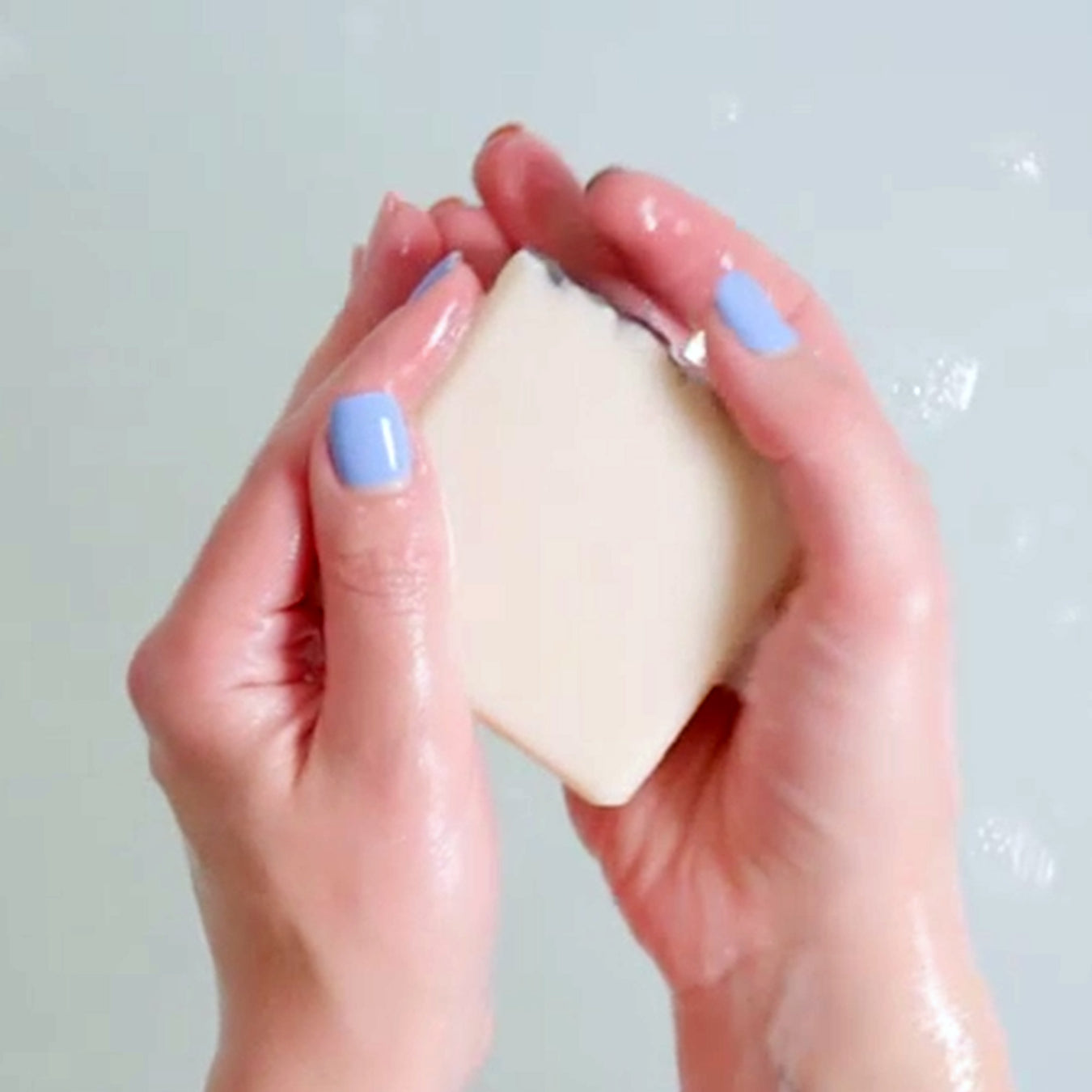 A pair of hands with blue nail polish carefully rub a soap bar in water.