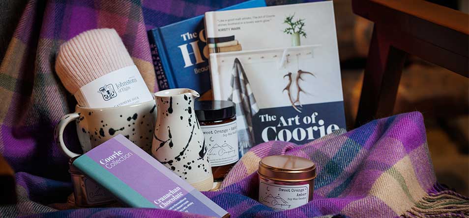 Collection of Coorie products including candles, chocolate, cashmere socks and tartan throws are placed together in front of a warming room. 