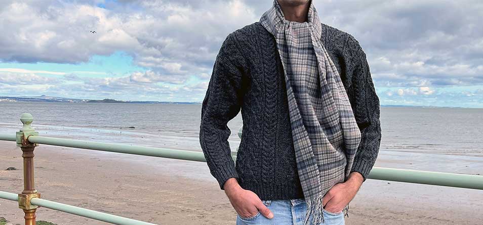 Person stands against a green beach railing wearing a charcoal aran jumper and tartan scarf. 