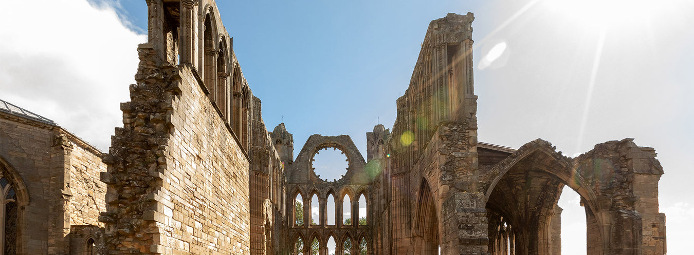 Part of Elgin Cathedral ruins against a bright blue sky with sunbeams flooding the central wall. 