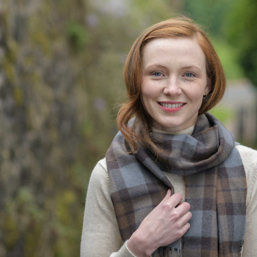 A person stands next to a brick wall wearing the Outlander wool Scarf. The person smiles whilst holding the scarf close to their chest. 