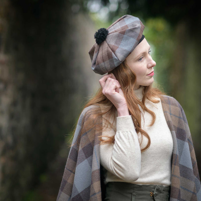 Person stands next to a brick wall wearing the Outlander Tartan Tam and Serape. The tartan features a brown and blue check. 