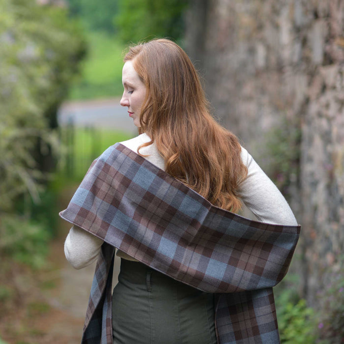 Person standing on a bricked path with their back to the camera wearing the Outlander Tartan Sash across their shoulders. The Sash features a brown and blue check.