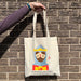 A person holds up a cotton tote bag against a brick wall. The tote is a natural colour with a colourful depiction of Robert the Bruce. 
