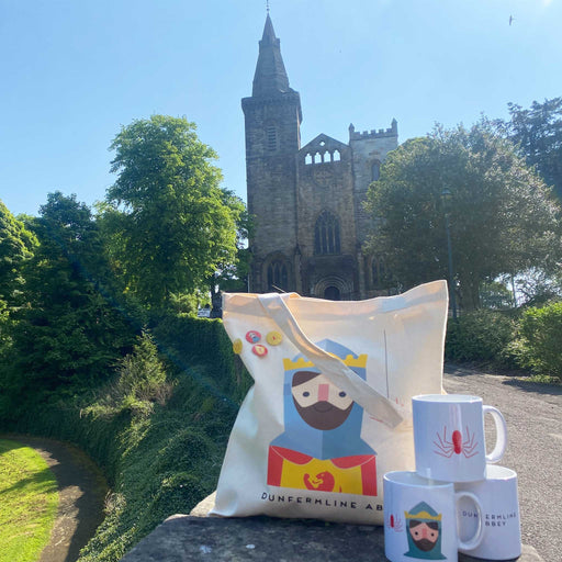 a set of 3 Robert the Bruce mugs are displayed on a wall with a tote bag. Dunfermline Abbey is in the background.  