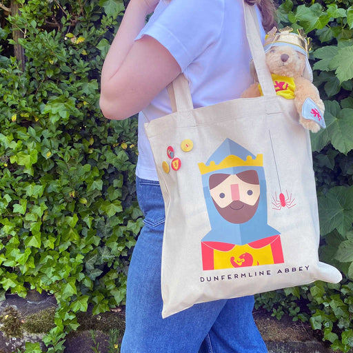 A person wears over their shoulder the cotton Robert the Brice tote. A Robert the Bruce teddy is popping out of the top of the bag. 